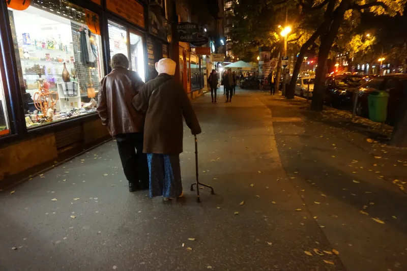 An elderly couple walking arm in arm along a shop-lit street at dusk, the woman with a cane