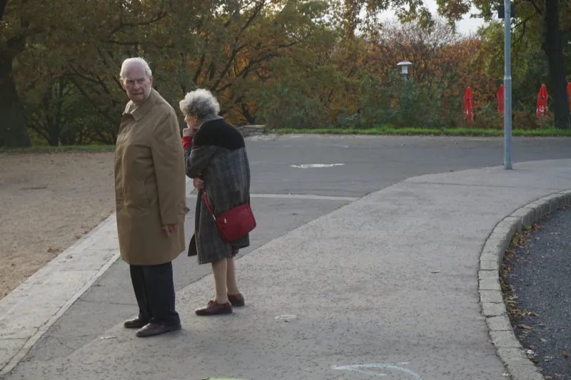 An elderly couple standing on a curving path in an autumn park, the woman holding a red handbag