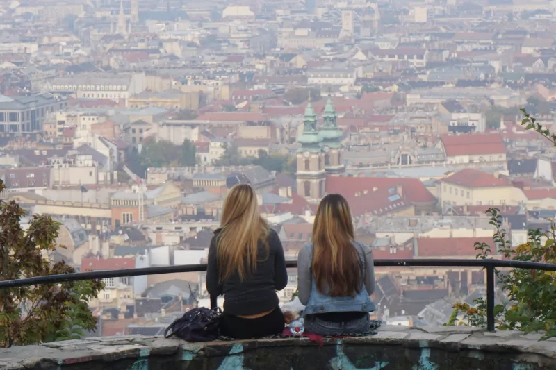 Two young women with long hair sitting on a wall, looking out over the rooftops of a city