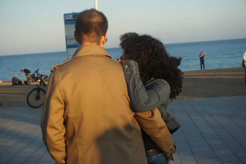 A man in a tan coat with his arm around a curly-haired woman, looking out at the sea