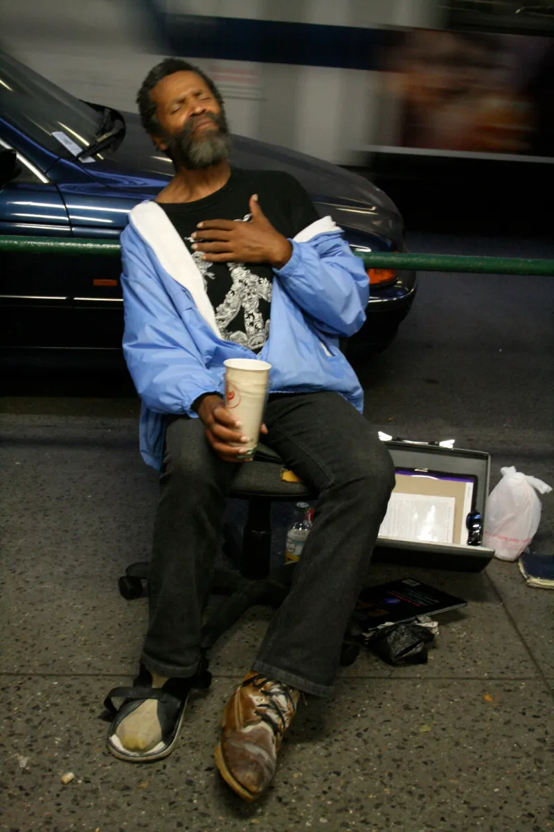 A man seated on a city sidewalk at night with his eyes closed, a hand on his chest and a paper cup in the other