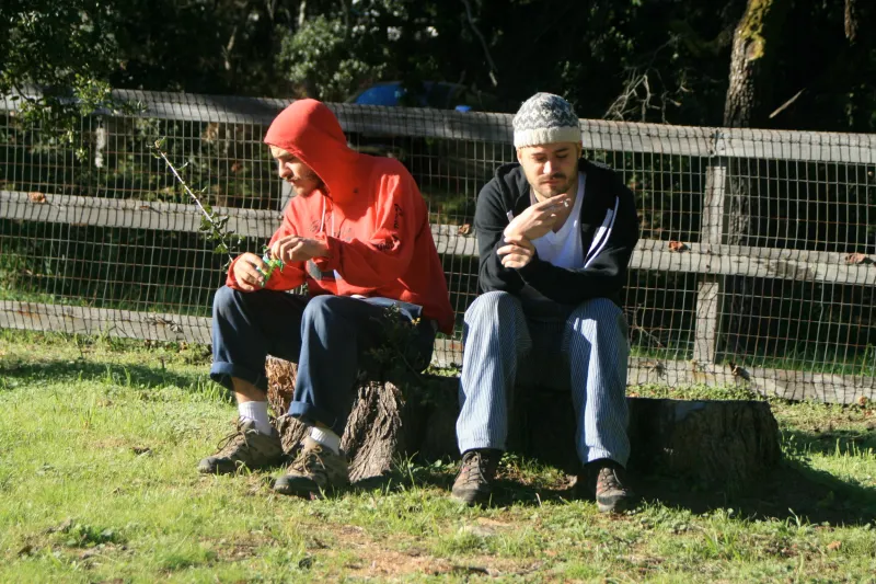 Two men sitting on a log in the sun, eating, one in a red hoodie