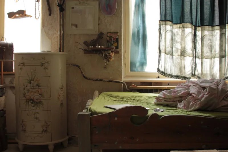 A small bedroom with a painted chest of drawers and an unmade bed by a curtained window