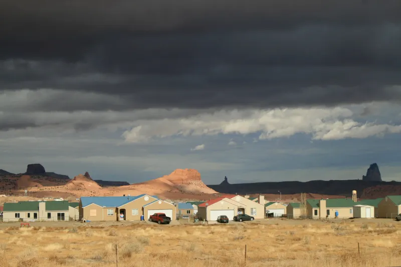 A row of houses on a plain beneath red mesas and storm clouds