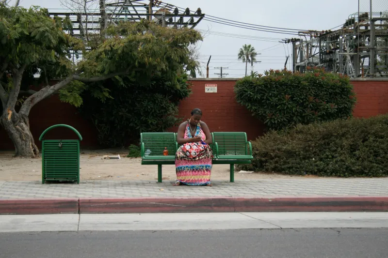 A woman in a colourful striped dress sitting alone on a green bench by a quiet road