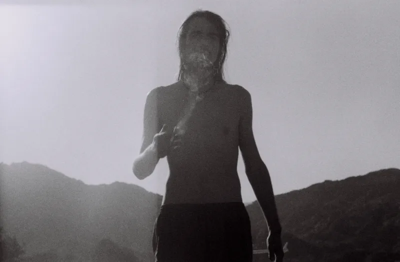 A shirtless woman smoking against hazy mountains, in black and white