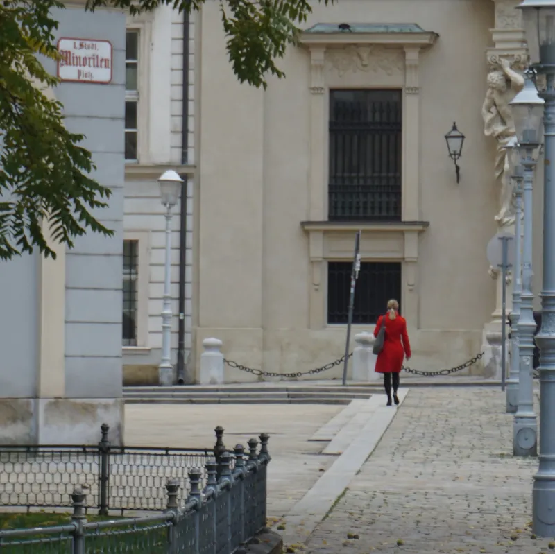 A woman in a red coat walking past a grand pale building on a cobbled street