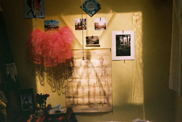 A room wall hung with a red tutu, photographs and a skeleton poster, in warm light